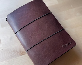 May include: A dark brown leather journal cover featuring two thin black elastic bands wrapped horizontally around the exterior. The word Chickee is embossed in the bottom right corner. Silver metal eyelets are visible along the spine edge.