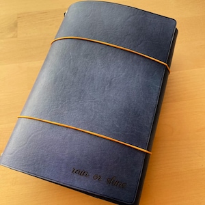 May include: A dark blue notebook with a textured cover secured by two thin orange elastic bands. The bottom right corner of the cover features the embossed text "rain or shine" in a cursive script font. The notebook has a rectangular shape.