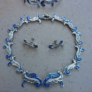 Puede incluir: Un conjunto de collar, pulsera y pendientes de plata con esmalte azul. Las joyas presentan un diseño floral delicado.