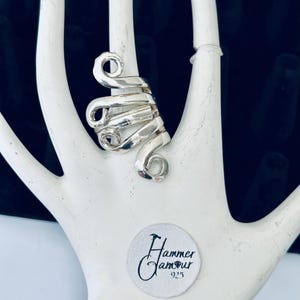 May include: A silver-colored ring with a spiral design. The ring is wrapped around a white hand-shaped display. The ring has a hammered texture and is marked with the text "Hammer Glamour 925" on a white sticker.