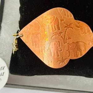 May include: A copper-colored bracelet with an elephant design. The bracelet features a heart-shaped pendant with etched elephants and palm trees. The pendant is connected to a silver chain. A small gold charm is attached to the chain.