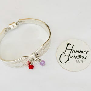 May include: A silver bracelet crafted from a spoon, featuring intricate floral patterns. The bracelet has a magnetic clasp and is adorned with red, clear, and purple crystal charms. A circular tag with the text "Hammer Glamour 925" is also visible.