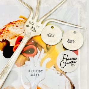May include: A silver fork with four tines, engraved with "Hair of the Dog" and a sleeping dog illustration. Includes three small round tags with dog illustrations and a tag with "Hammer Glamour 925" text. The background shows a Bloody Mary.