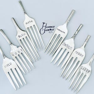 May include: A set of eight silver forks with different cheese names engraved on them, including Brie, Bleu, Goat, Mild, Cheddar, Swiss, Colby, and Sharp. The forks are arranged in a circular pattern on a white background.