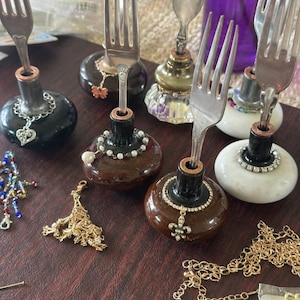 May include: A collection of handcrafted fork holders, each featuring a repurposed fork inserted into a decorative base. The bases vary in color and design, including black, brown, and white, adorned with beads and charms. A variety of jewelry pieces are scattered around.