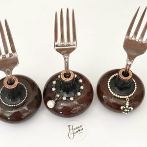 May include: Three decorative fork holders. Each holder features a vintage fork attached to a dark brown, rounded base. One has a pearl necklace, another a fleur-de-lis pendant, and the third has a ring of small crystals.