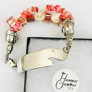 May include: A silver bracelet with a curved, blank nameplate and a beaded band. The band features pink and white beads, with silver floral accents at each end. The bracelet is displayed on a white surface.