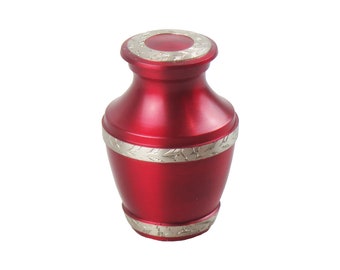 Op de afbeelding: Een kleine rode metalen urn met zilveren versieringen en bloemmotieven. De urn heeft een ronde bovenkant en een licht uitlopende basis.