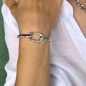 May include: A silver-colored cuff bracelet with a horseshoe-shaped design. The bracelet is worn on a wrist, and the background includes a white shirt and a green surface.