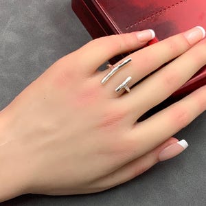 May include: A silver-colored ring with a modern design. The ring features two straight, bar-shaped elements that wrap around the finger. The ring is displayed on a hand with manicured nails.