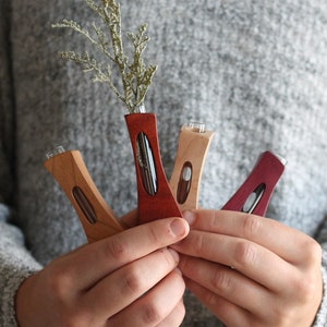 May include: Four wooden bud vases in different colors and wood grains. The vases are held in a person's hands. The vases have a clear glass tube inside for holding flowers or greenery.