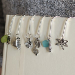 May include: Seven silver bookmark clips with different charms, including a dragonfly, a musical note, a bee, a small house, a green glass bead, and a blue glass bead.