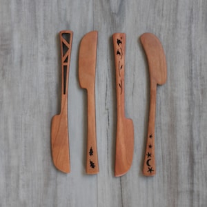 May include: Four wooden butter knives with different carved designs. The knives are all made of light brown wood and have a slightly curved blade. The designs include a geometric pattern, a tree, a vine, and stars and a crescent moon.