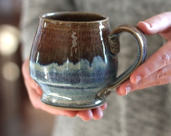 Może przedstawiać: Kubek ceramiczny w kolorze brązowym i niebieskim, o szerokim, zaokrąglonym korpusie i uchwycie. Kubek ma szkliwo, które tworzy marmurkowy efekt, z brązowymi i niebieskimi odcieniami.