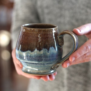 May include: A brown and blue ceramic mug with a wide, rounded body and a handle. The mug has a glaze that creates a mottled effect, with brown and blue shades.