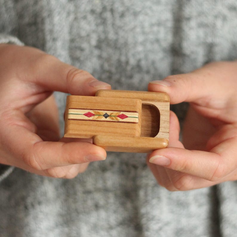 May include: A small rectangular wooden box with a sliding lid. The lid features a decorative inlay strip with a pattern of red diamonds and a central black square, set against light wood grain. The box has a hollow compartment on the right side.