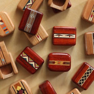 May include: A collection of rectangular wooden blocks featuring polished surfaces and intricate inlaid geometric patterns in various colors including red, brown, tan, black, blue, and yellow. Each block displays unique decorative wood marquetry.