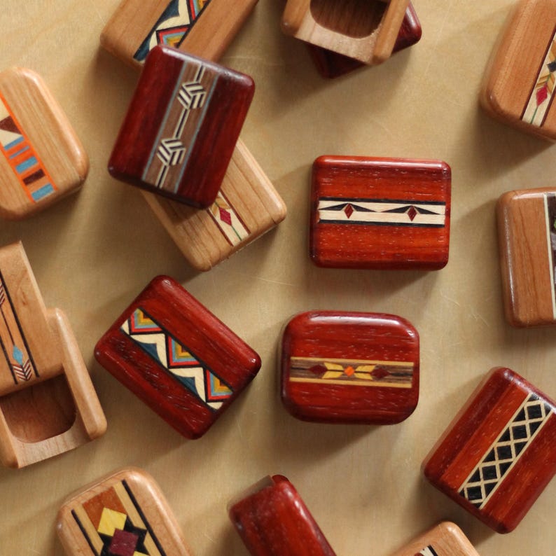 May include: A collection of rectangular wooden blocks featuring polished surfaces and intricate inlaid geometric patterns in various colors including red, brown, tan, black, blue, and yellow. Each block displays unique decorative wood marquetry.