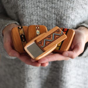 May include: A collection of small, wooden compact mirrors with intricate geometric designs in shades of brown, red, blue, and yellow. One mirror is open, revealing a reflective surface. The mirrors are held in open hands against a gray background.