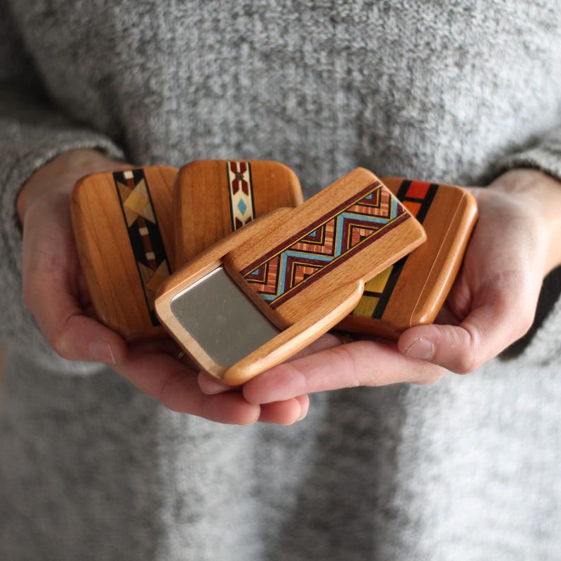 May include: A collection of small, wooden compact mirrors with intricate geometric designs in shades of brown, red, blue, and yellow. One mirror is open, revealing a reflective surface. The mirrors are held in open hands against a gray background.