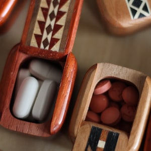 May include: Two rectangular wooden pill boxes with hinged lids. The left box is reddish-brown with a geometric inlay pattern and contains white oval tablets. The right box is light brown with a dark inlay and contains small round orange tablets.