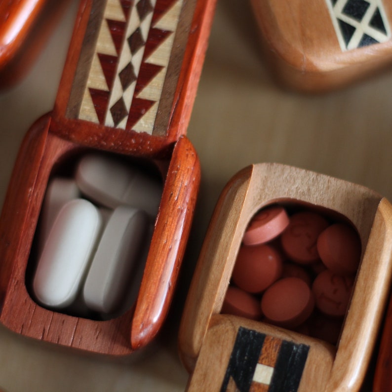 May include: Two rectangular wooden pill boxes with hinged lids. The left box is reddish-brown with a geometric inlay pattern and contains white oval tablets. The right box is light brown with a dark inlay and contains small round orange tablets.