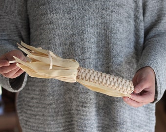 May include: A hand holds a woven corn husk decoration, resembling a corn cob. The husk is a light beige color and has a natural, rustic look.