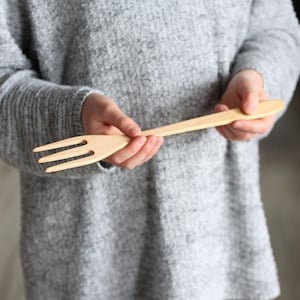 May include: A person is holding a wooden fork with a natural finish. The fork is long and has four tines.