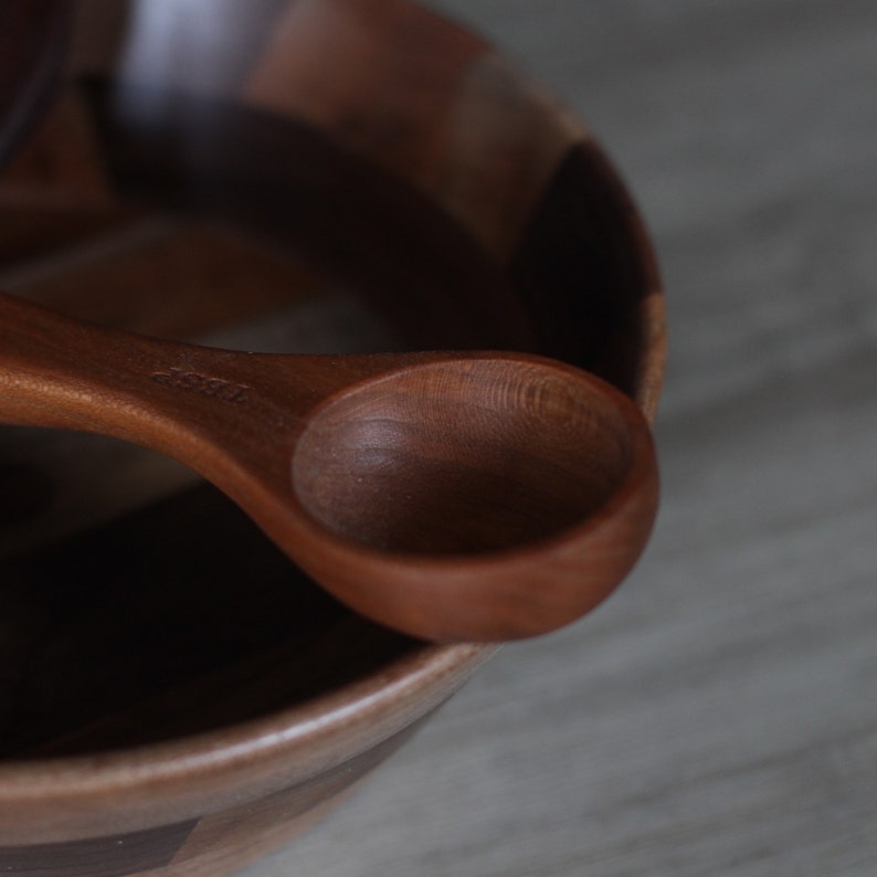 May include: A wooden bowl with a natural wood grain pattern and a wooden spoon resting on the rim. The spoon has a rounded bowl and a handle with the word "1/4 cup" engraved on it.
