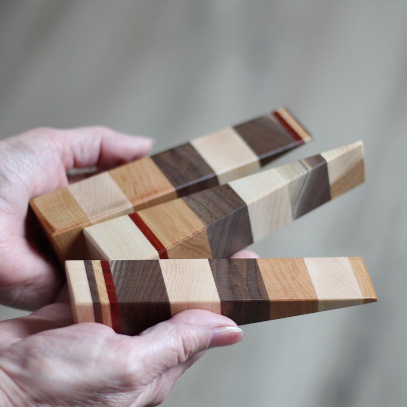Puede incluir: Tres topes de puerta de madera con un patr&oacute;n de rayas multicolores. Los topes de puerta est&aacute;n hechos de diferentes tipos de madera, incluyendo nogal, arce y cerezo. Los topes de puerta se sostienen en las manos de una persona.