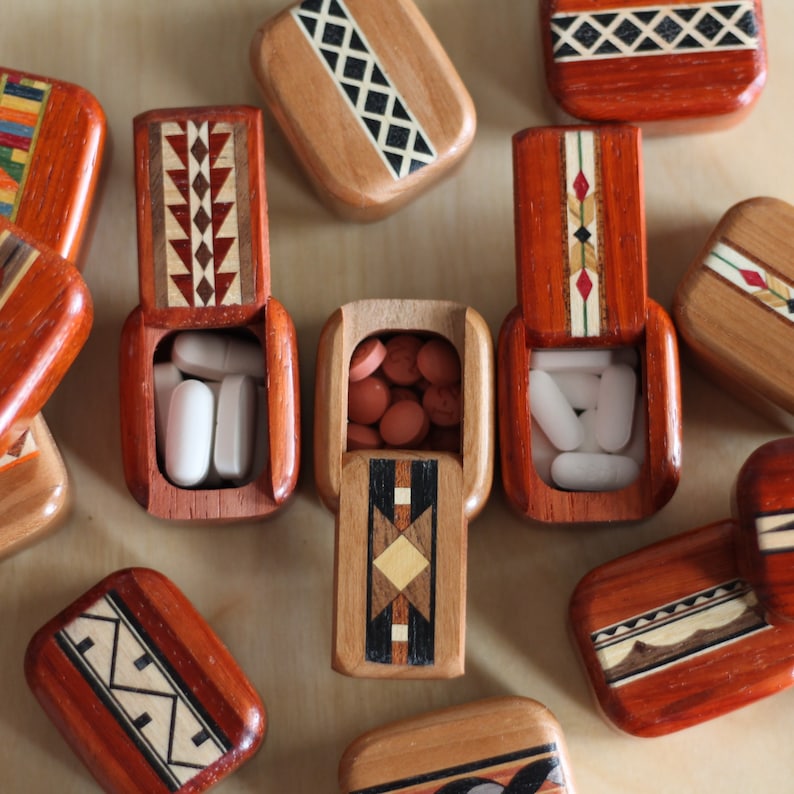 May include: A collection of small rectangular wooden pill boxes with hinged lids featuring inlaid geometric patterns in various wood tones. Some boxes are open, revealing white or reddish-brown round and oval tablets inside the compartments.