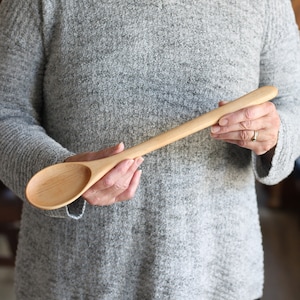 May include: A long, wooden spoon with a natural finish. The spoon is held by a person's hands.