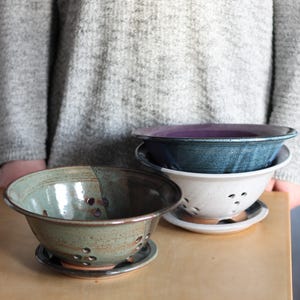 May include: Three ceramic colanders with drainage holes. The colanders are stacked on top of each other. The top colander is purple, the middle colander is blue, and the bottom colander is white.