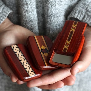 May include: Three small, rectangular wooden boxes with intricate inlaid designs. The boxes are primarily reddish-brown with accents of lighter wood and geometric patterns. One box is open, revealing a small mirror.