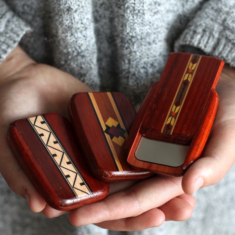 May include: Three small, rectangular wooden boxes with intricate inlaid designs. The boxes are primarily reddish-brown with accents of lighter wood and geometric patterns. One box is open, revealing a small mirror.