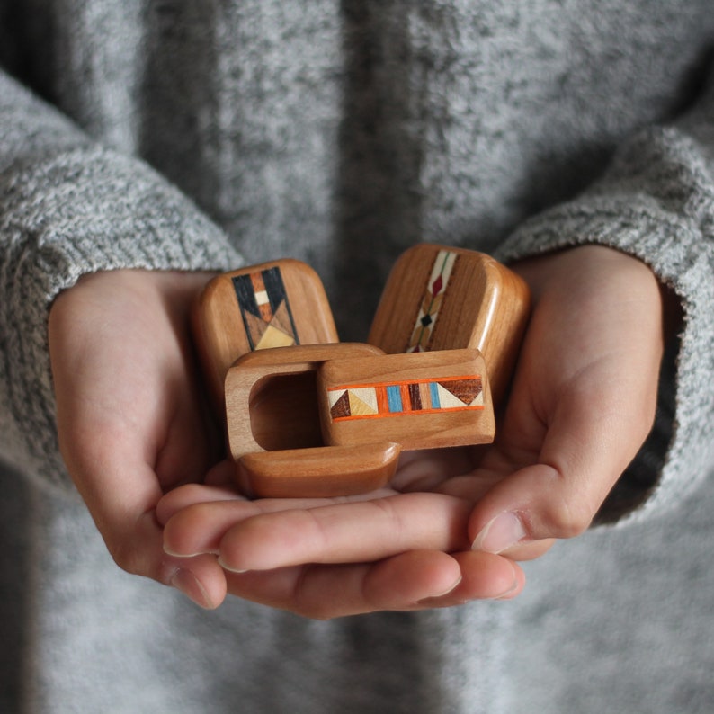 May include: Three small wooden boxes with rounded corners and smooth surfaces. Each box features a unique inlaid geometric pattern on the lid, composed of various light and dark wood tones, including orange, blue, and cream accents.