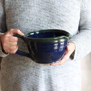 May include: A large blue ceramic mixing bowl with a green rim and a handle. The bowl is being held by a person's hands.