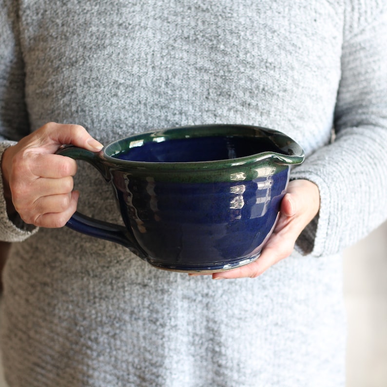May include: A large blue ceramic mixing bowl with a green rim and a handle. The bowl is being held by a person's hands.