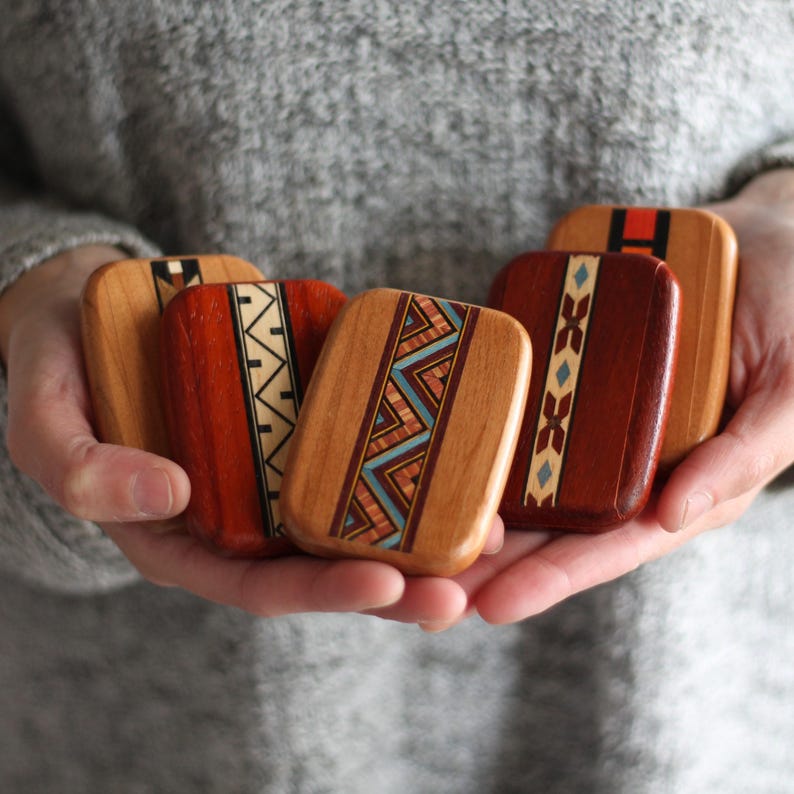 May include: A collection of rectangular wooden boxes with intricate inlaid designs. The boxes feature a variety of colors, including natural wood tones, red, and accents of blue and white. The boxes are held in a person's hands.