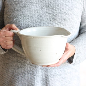 May include: A large white ceramic mixing bowl with a handle. The bowl has a speckled glaze and a slightly rounded shape.