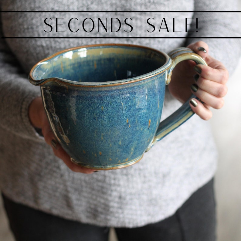 May include: A large, blue ceramic mixing bowl with a handle. The bowl has a speckled glaze with a light brown rim. The bowl is being held by a person's hands.
