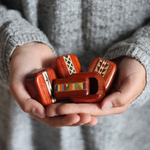 May include: A collection of four small, polished reddish-brown wooden objects with inlaid geometric patterns. Each piece features distinct decorative details, including cross-hatching, triangular motifs, and a central rectangular inlay with multicolored segments.