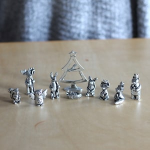 May include: A set of 10 silver-toned pewter figurines depicting woodland animals and a Christmas tree. The figurines include a moose, owl, rabbit, squirrel, raccoon, fox, and a bear. The set also includes a small bird perched on the Christmas tree.