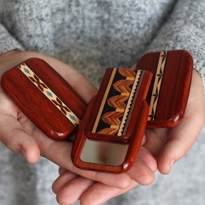 May include: Three rectangular wooden compact mirrors with intricate inlaid designs. The mirrors are a rich reddish-brown color, with decorative patterns in various wood tones and accents of blue. One mirror is open, revealing a reflective surface.