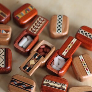 May include: A collection of small, rectangular wooden pill boxes with hinged lids. Each box features unique, intricate geometric inlay patterns in various wood tones. Some boxes are open, revealing white or reddish-brown oval-shaped pills inside.