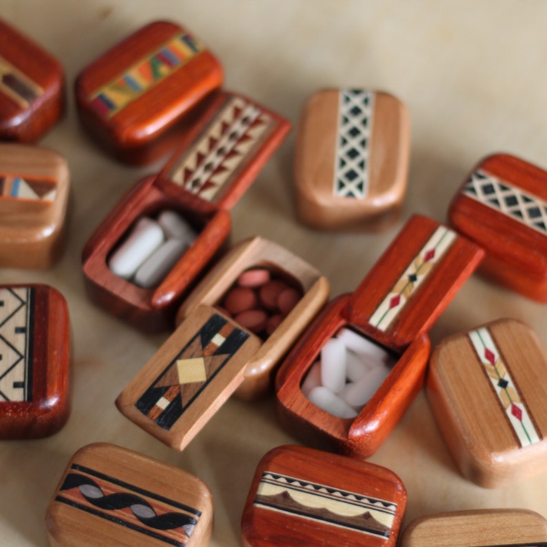 May include: A collection of small, rectangular wooden pill boxes with hinged lids. Each box features unique, intricate geometric inlay patterns in various wood tones. Some boxes are open, revealing white or reddish-brown oval-shaped pills inside.