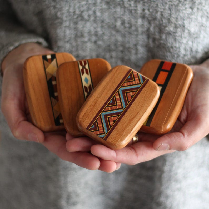 May include: Four rectangular wooden boxes with inlaid geometric designs in various colors are held in open hands. The boxes have a warm wood tone and are polished. The designs include triangles, zigzags, and rectangular patterns.