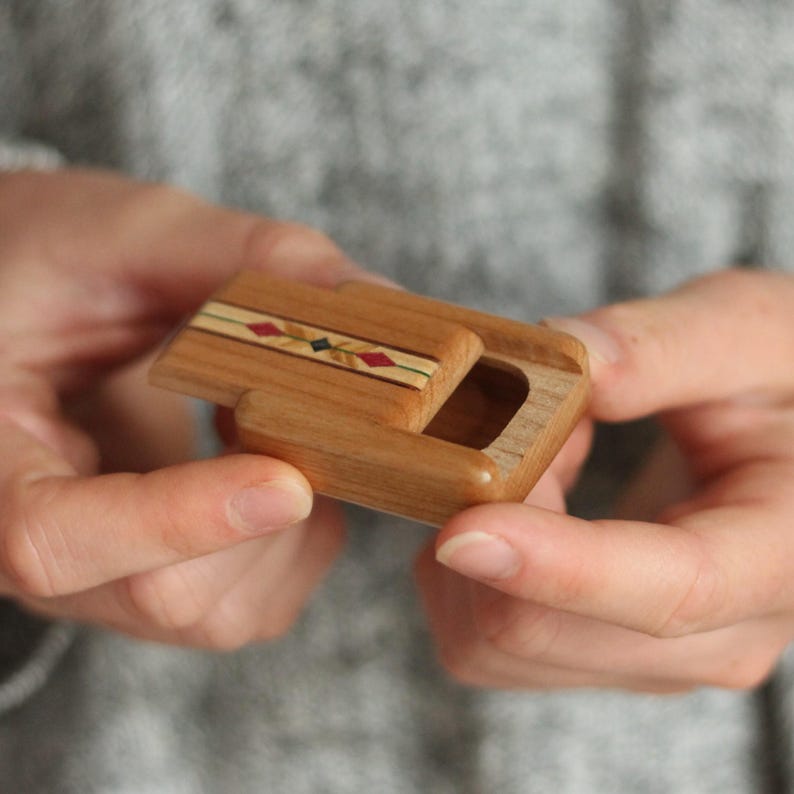 May include: A small rectangular wooden box with a sliding lid. The lid features a decorative inlay strip with a repeating pattern of red and dark green diamond shapes set against a light tan background, contrasting with the natural brown wood grain.