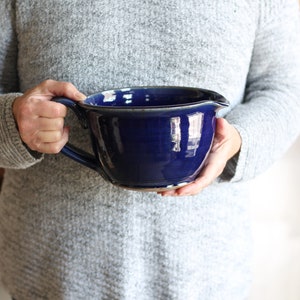 May include: A large, deep blue ceramic mixing bowl with a handle. The bowl has a glossy finish and a slight brown rim.