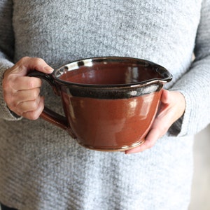 May include: A large, brown ceramic mixing bowl with a black rim and a handle. The bowl is glazed and has a rustic look.